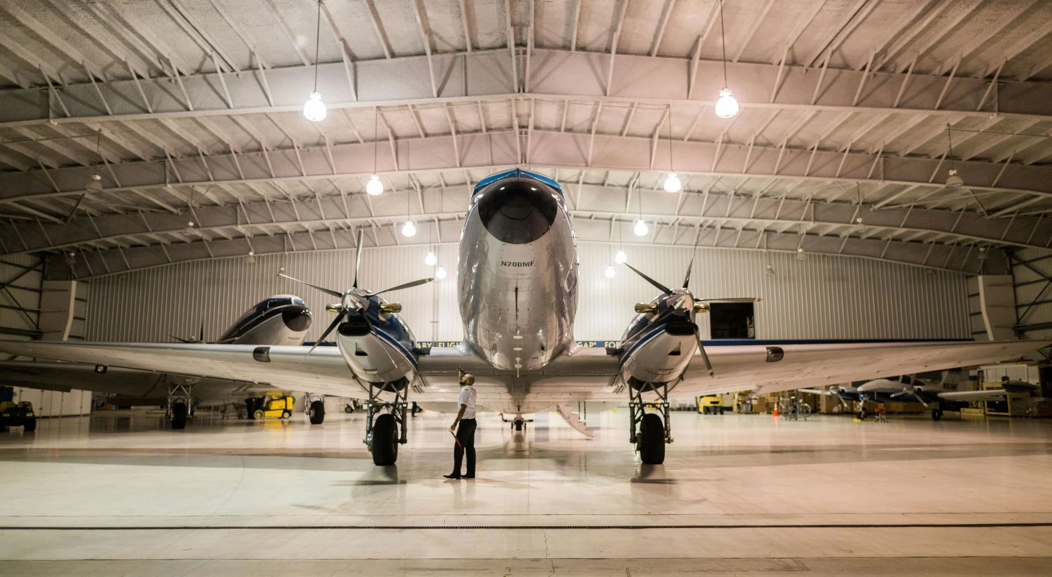 A man incpecting a plane parked in a hangar with bright lights above it. 