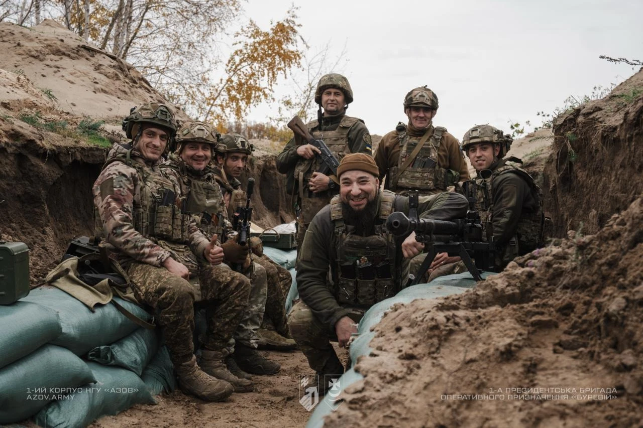 Група військовослужбовців посміхається, сидячи в окопі.