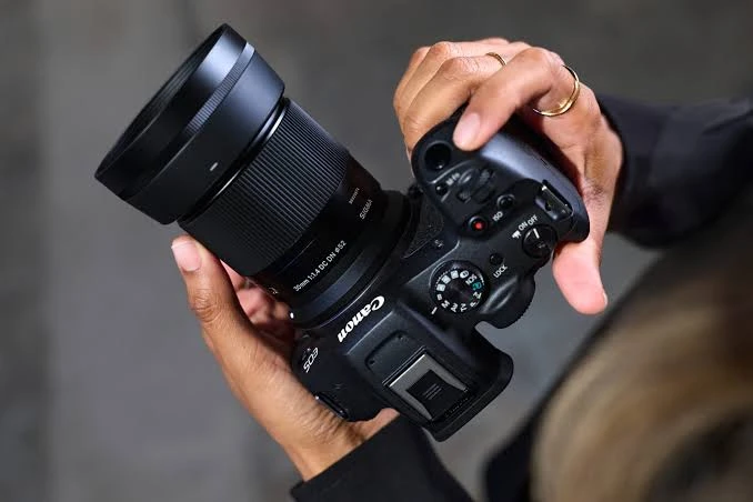 Руки тримають камеру Canon з встановленим об'єктивом Sigma 30mm 1.4 DC DN, великий палець лежить на кнопці спуску затвора, на оптику надіта масивна пластикова бленда.