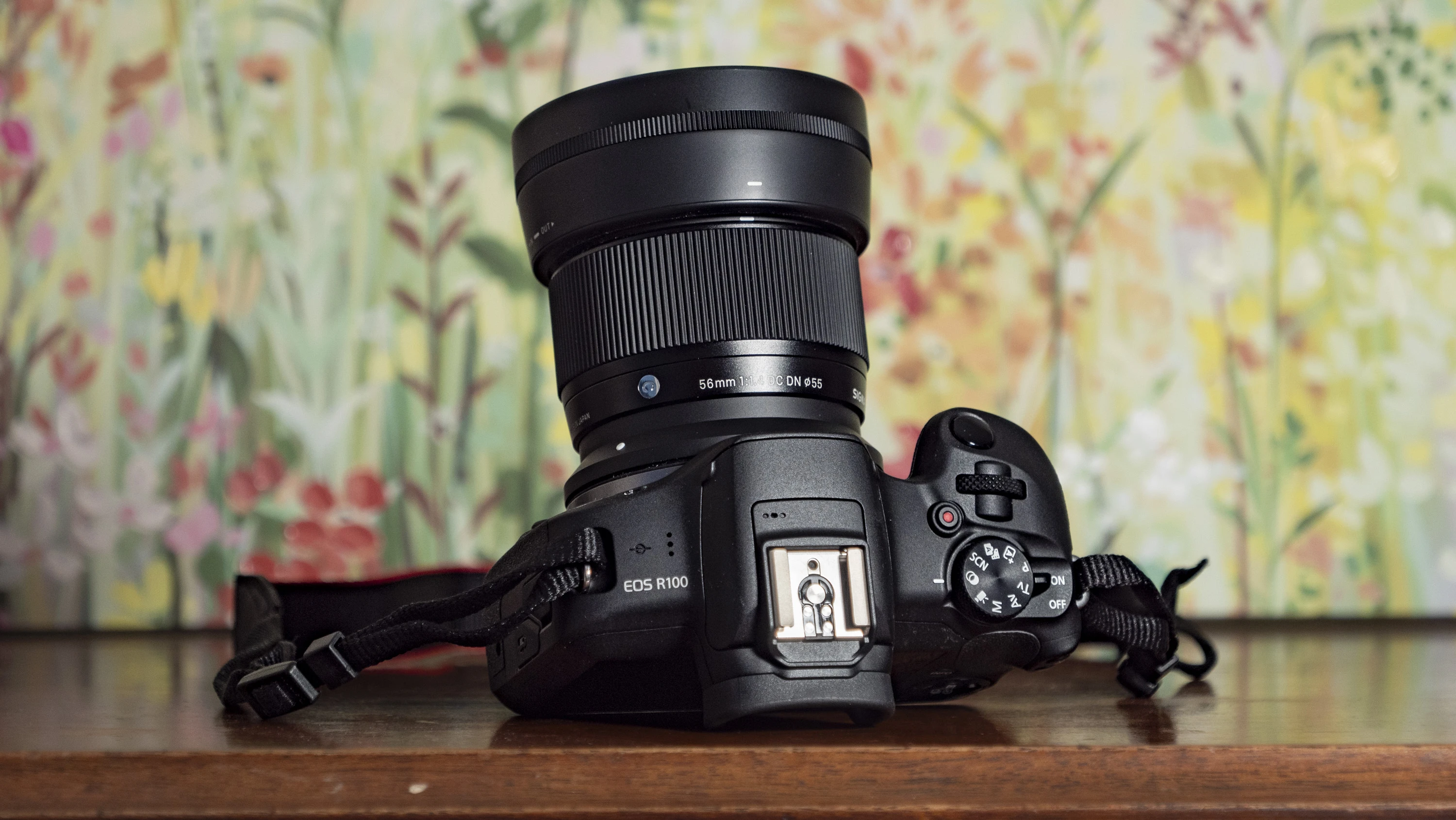 Портретний об'єктив Sigma 56mm 1.4 DC DN встановлений на компактну бездзеркальну камеру Canon EOS R100, яка лежить на дерев'яному столі на фоні світлих шпалер з рослинним візерунком.