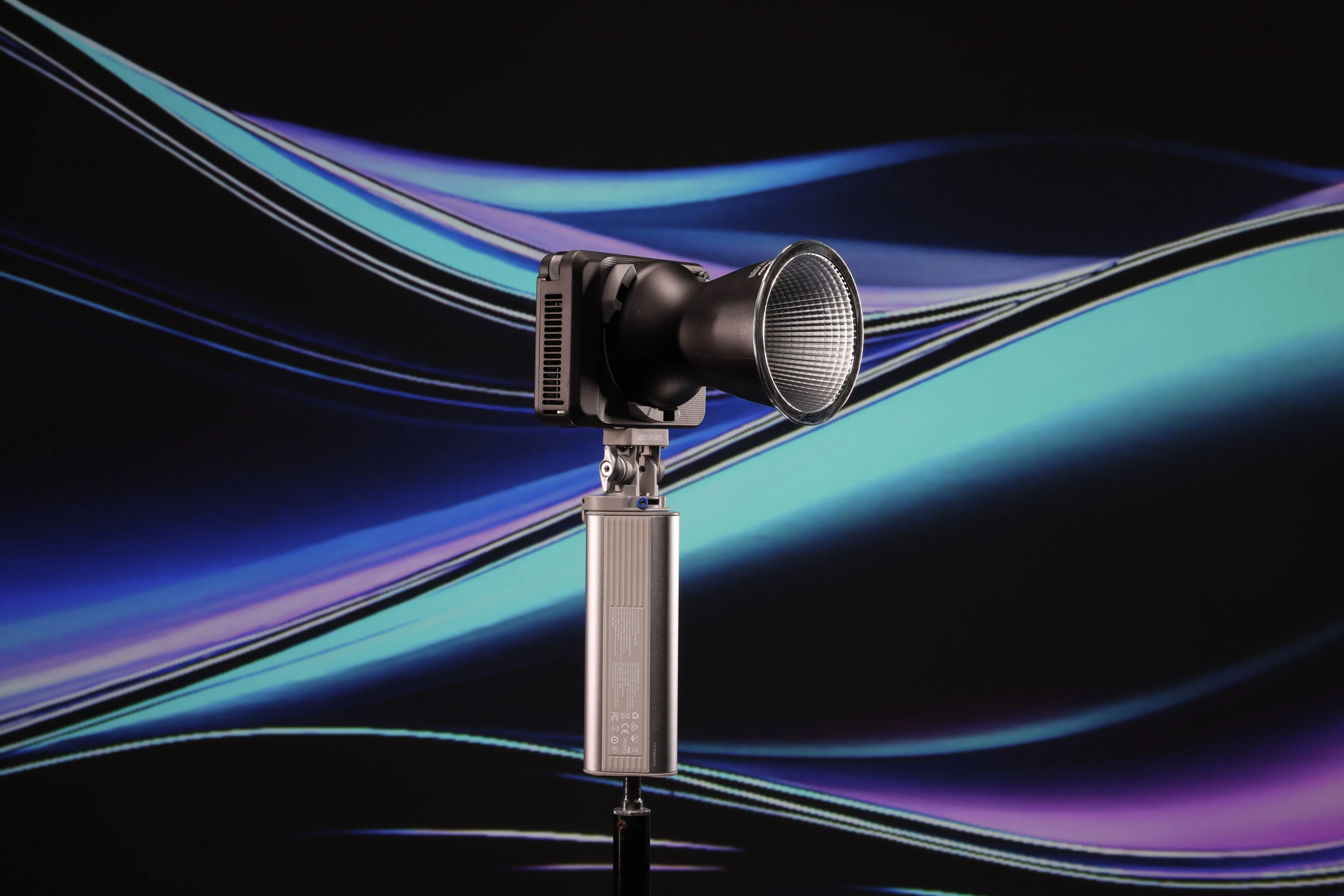 Компактний повнокольоровий RGB LED моноблок amaran Ray 120c з рефлектором, встановлений на акумуляторну рукоятку amaran Peak проти неонового тла