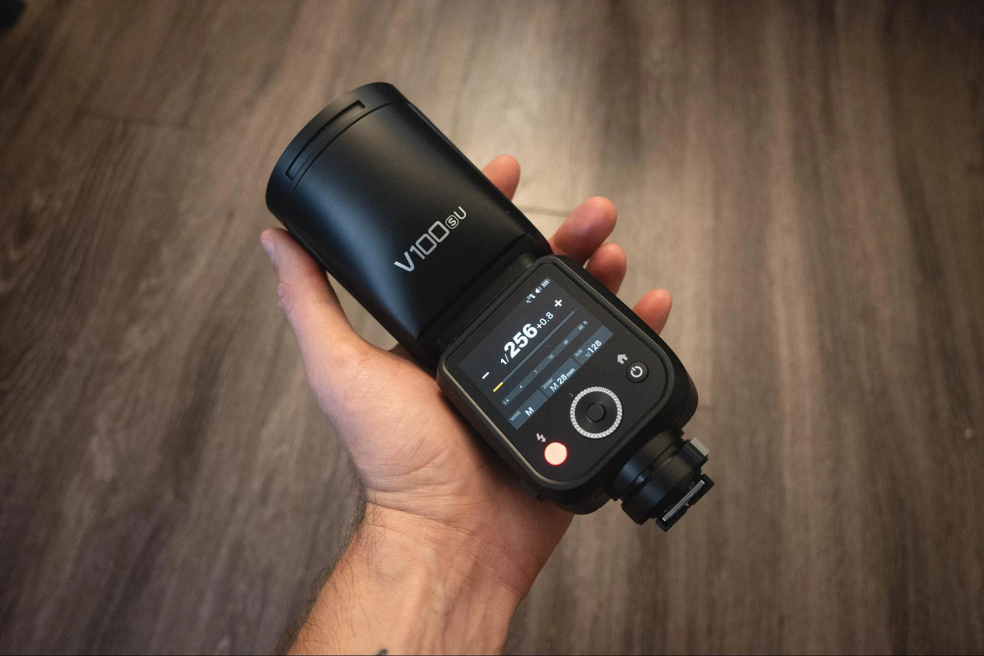 Спалах Godox V100 тримають у руці, на кольоровому дисплеї відображається потужність 1/256, режим M, зум 28 мм та субспалах