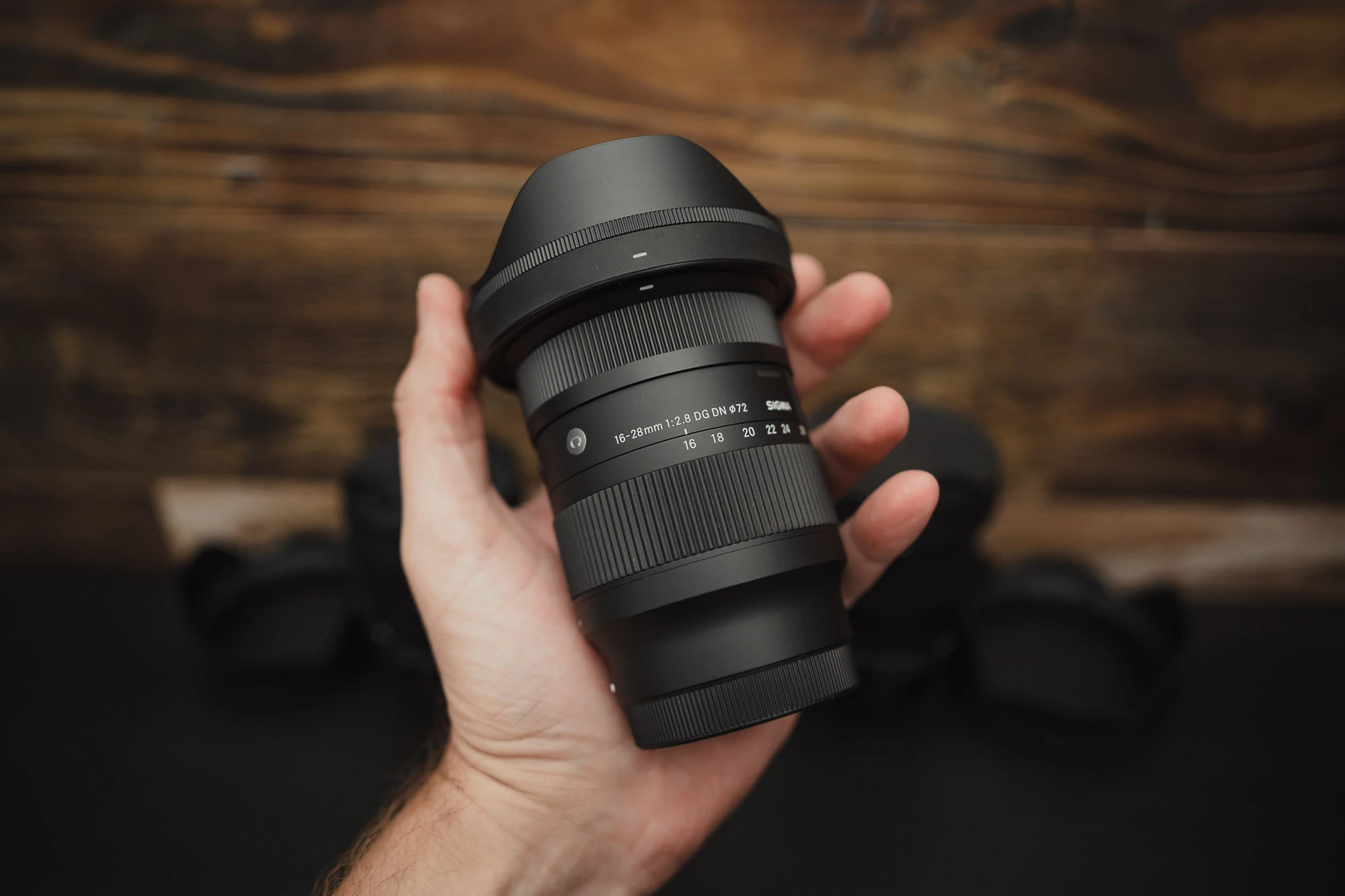 Компактний об'єктив Sigma 16-28mm f2.8 DG DN Contemporary у руці людини на тлі дерев'яної стіни