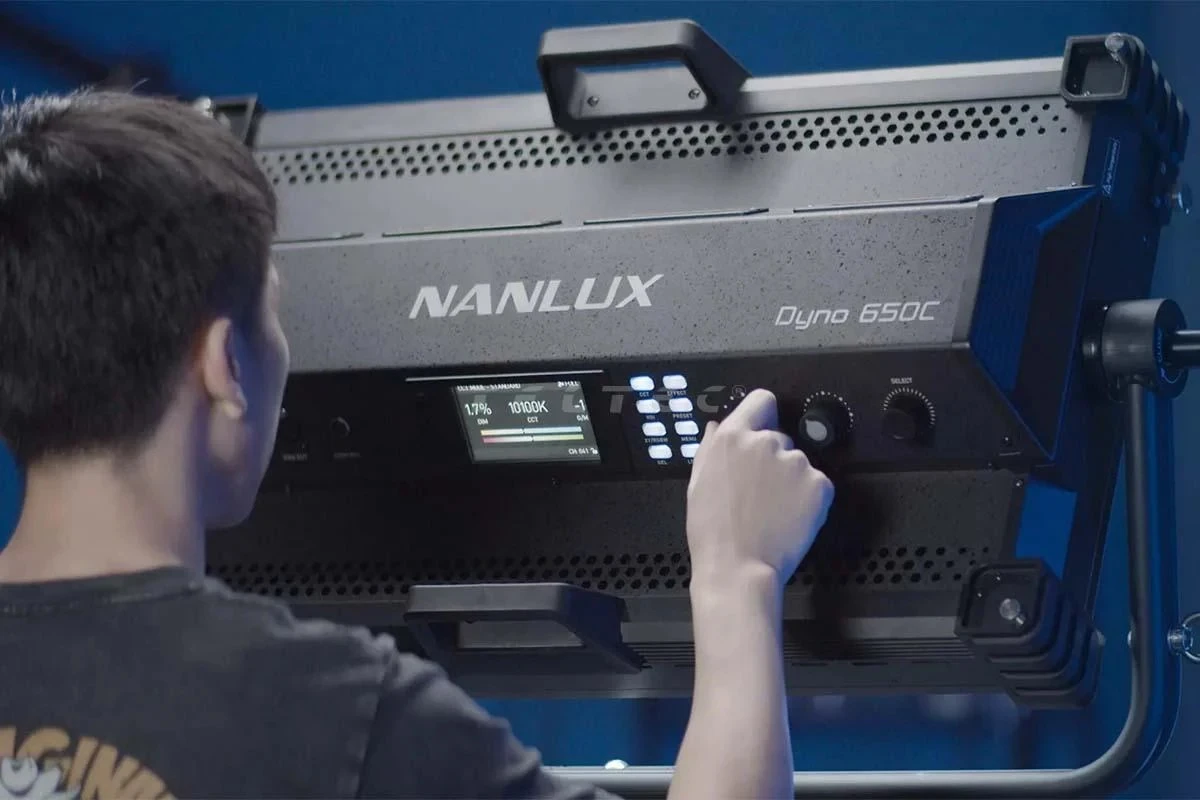 Задня панель кінопанелі Nanlux Dyno 650C у студійному напівмороці, де рука оператора налаштовує параметри CCT 10100K та корекцію G/M біля кольорового екрана