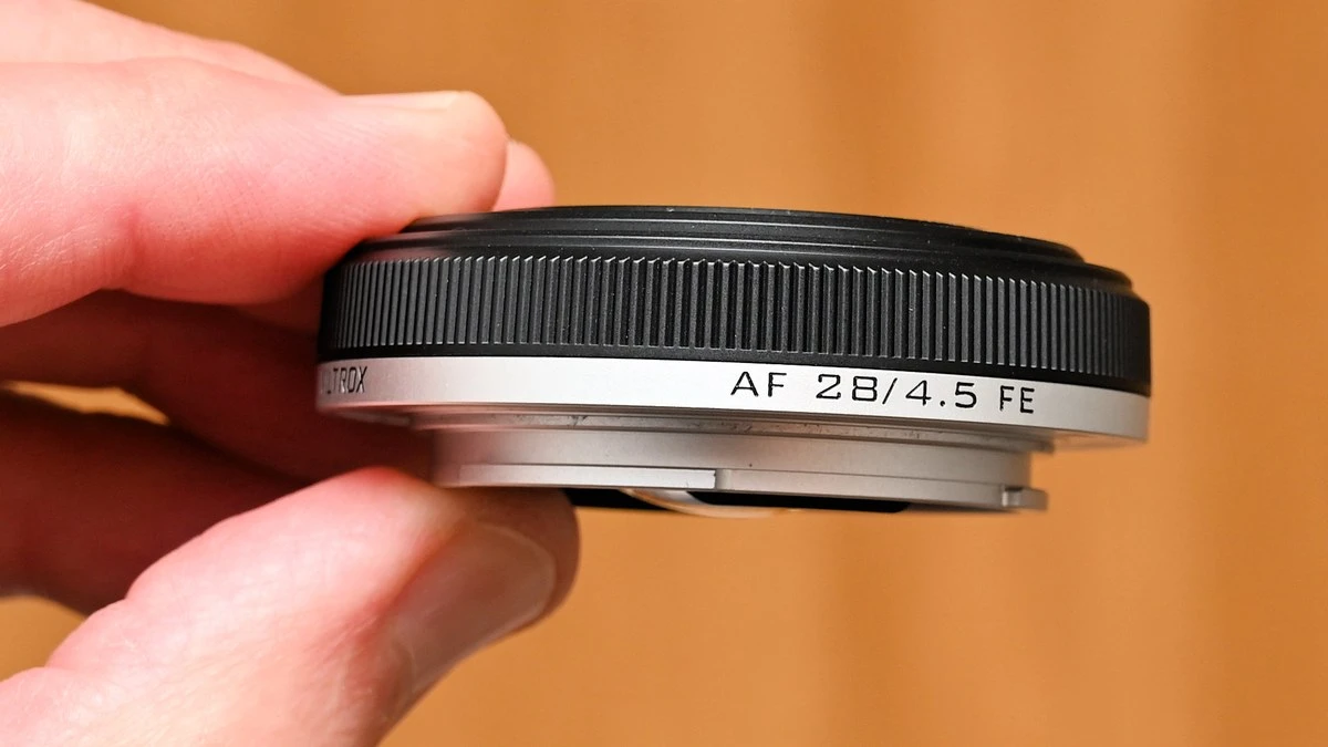 Объектив Viltrox AF 28mm f/4.5 FE толщиной 15.3 мм в руке, виден металлический байонет и серебристое кольцо с маркировкой.
