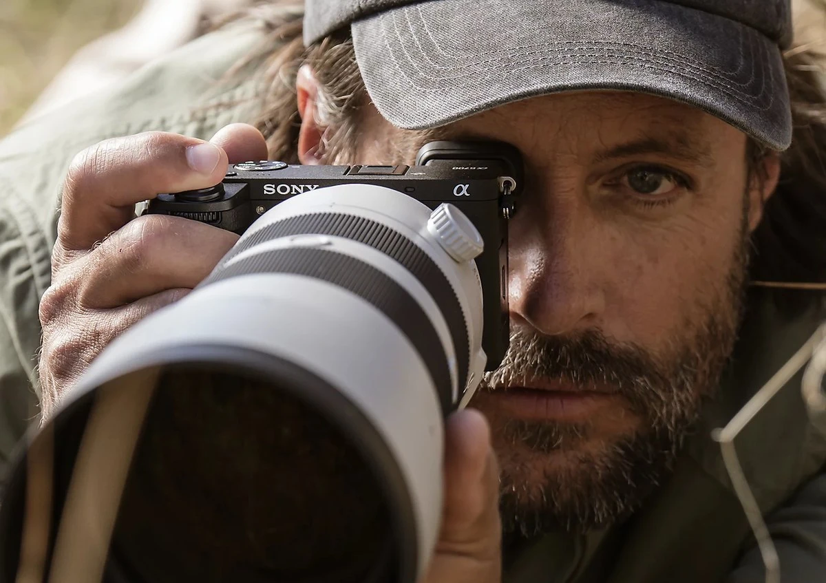 Фотограф в кепке визирует кадр через электронный OLED-видоискатель камеры Sony Alpha 6700 на природе