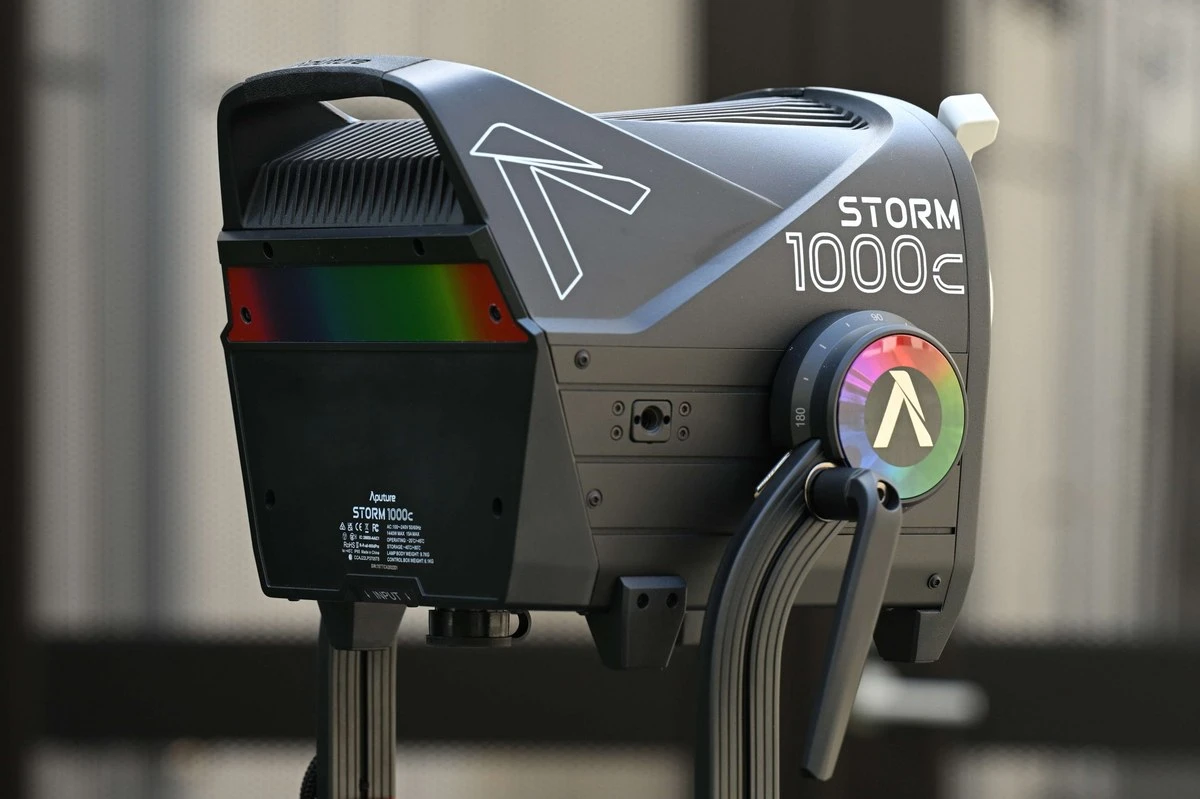 Моноблок Aputure STORM 1000c у ракурсі ззаду-збоку з райдужним диском ліри та роз'ємом живлення на індустріальному фоні