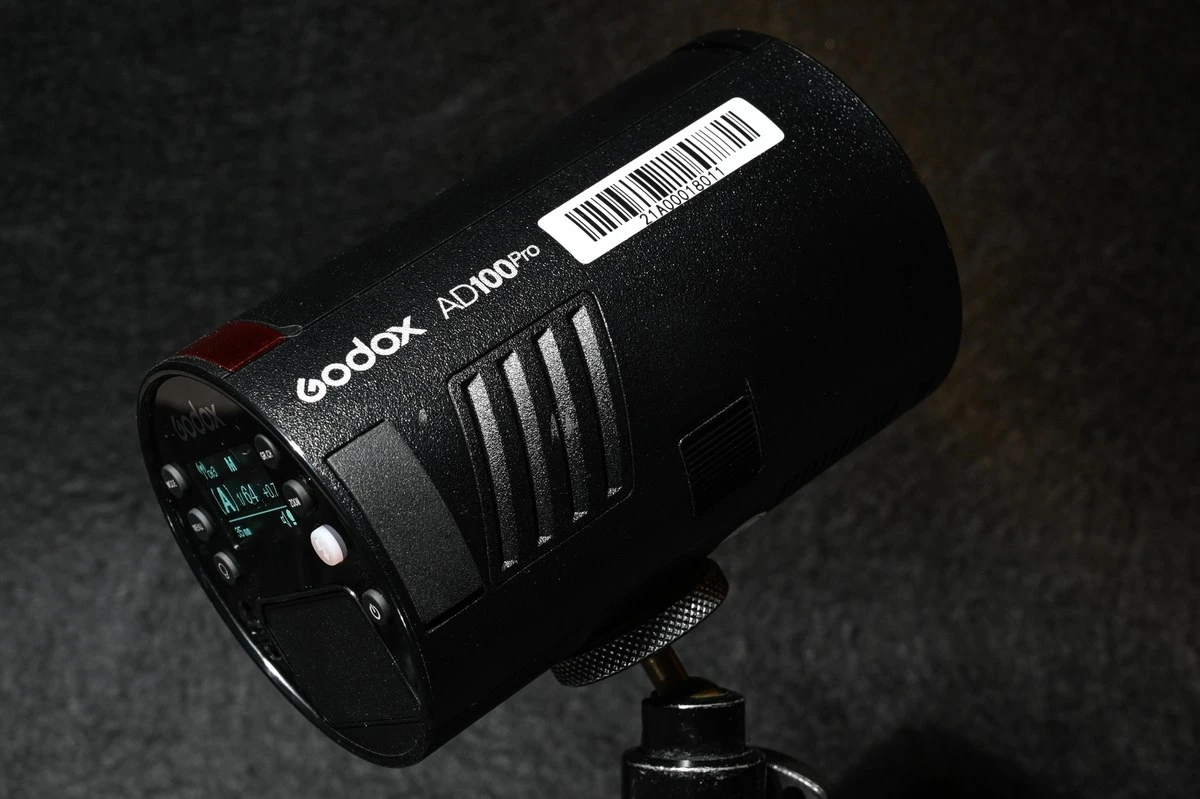 Компактний чорний циліндричний спалах Godox AD100Pro на металевому штативному адаптері з вентиляційними отворами, замком акумулятора та екраном CH3 M 1/64
