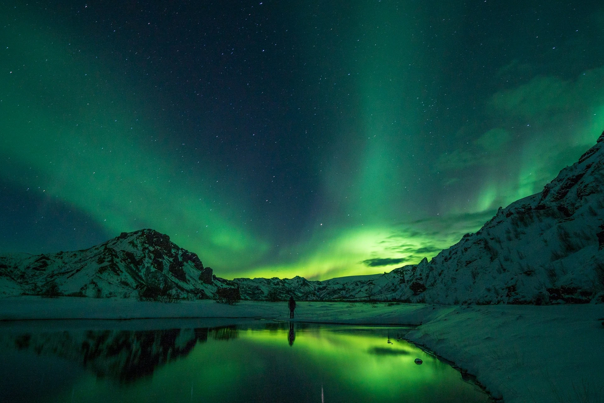 The Aurora Borealis dances above an individual standing near a lake as they take a photgraph.
