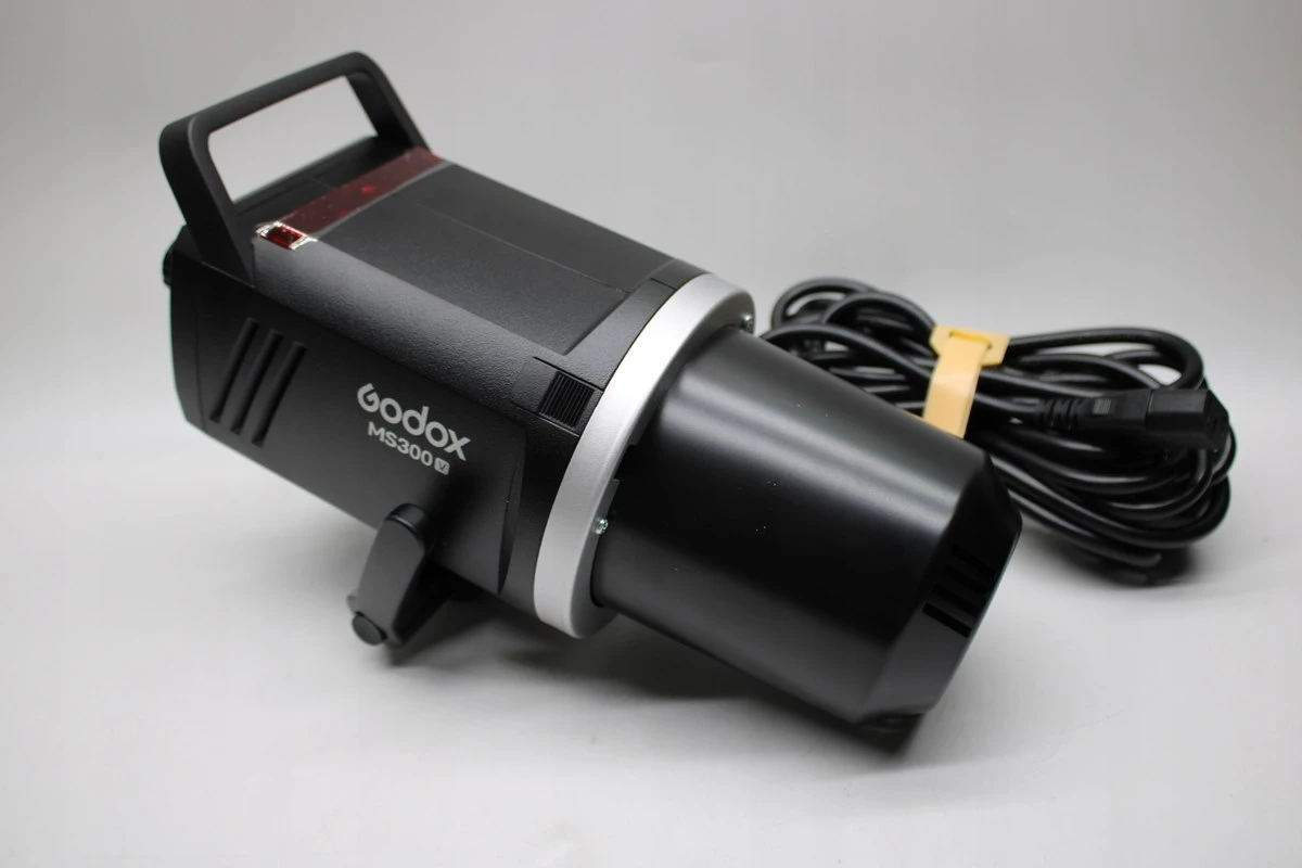Студійний моноблок Godox MS300-V на нейтральному столі у напівпрофіль зі встановленим захисним ковпаком, поруч лежить чорний мережевий кабель живлення із жовтою стяжкою
