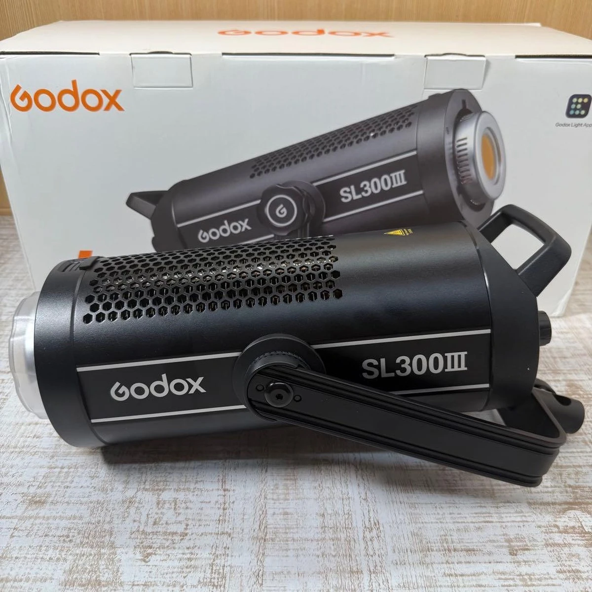 Студийный светодиодный моноблок Godox SL300III в горизонтальном положении на столе рядом с фирменной коробкой и металлической лирой крепления