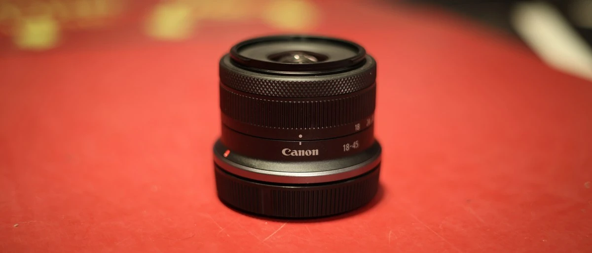 Крупный план ультракомпактного сложенного объектива Canon RF-S 18-45mm f/4.5-6.3 IS STM со снятой передней крышкой на ярко-красной поверхности стола