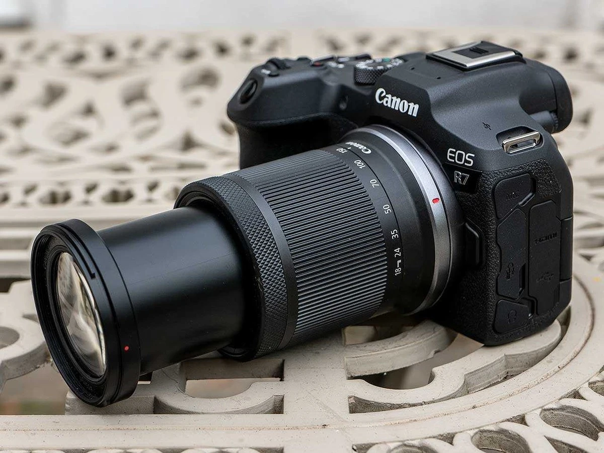 Камера Canon EOS R7 с установленным суперзумом Canon RF-S 18-150mm f/3.5-6.3 IS STM с максимально выдвинутым тубусом зума на белом металлическом садовом столике