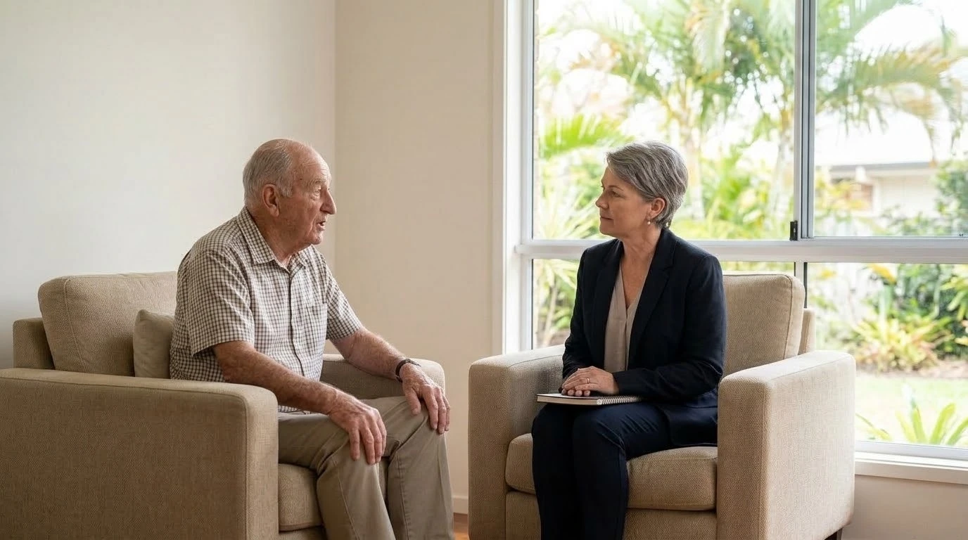 An older adult and a psychologist sit in two comfortable chairs in a calm consulting room, with space between them. The older person is speaking. The psychologist listens, notebook closed. Soft coastal light comes through a wide window, with a hint of palms outside, suggesting the Sunshine Coast without any signage. The right third of the frame is mostly window and light. Private, steady, and unsentimental. No couch cliché, no dark room, no hands covering a face. Aspect ratio 16:9.