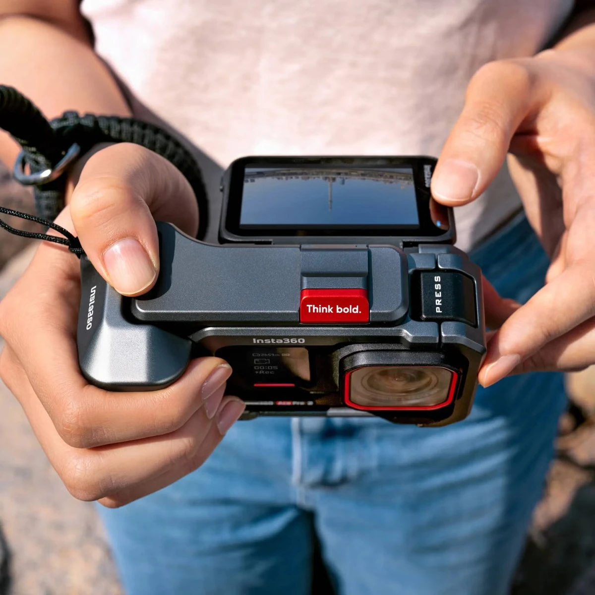 Описание: Камера Insta360 Ace Pro 2 у масивній захисній рамці-рукоятці Xplorer Grip Pro Kit з червоною клавішею спуску та ремінцем на руку, у руках оператора при денному світлі
