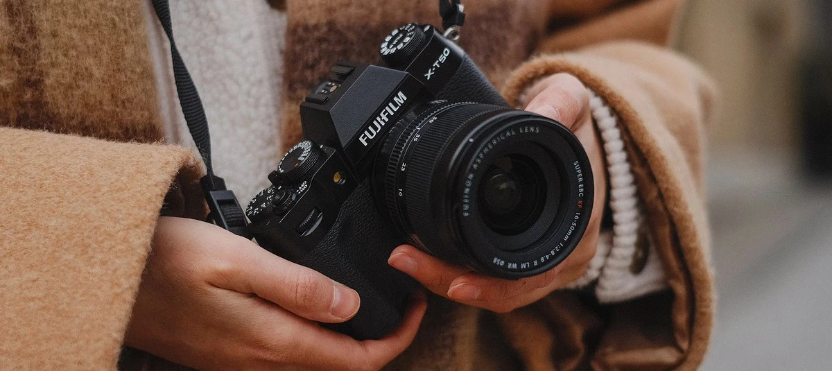 Людина в коричневому пальто тримає чорну камеру FUJIFILM X-T50 з зум-об'єктивом 16-50mm двома руками на нашийному ремені